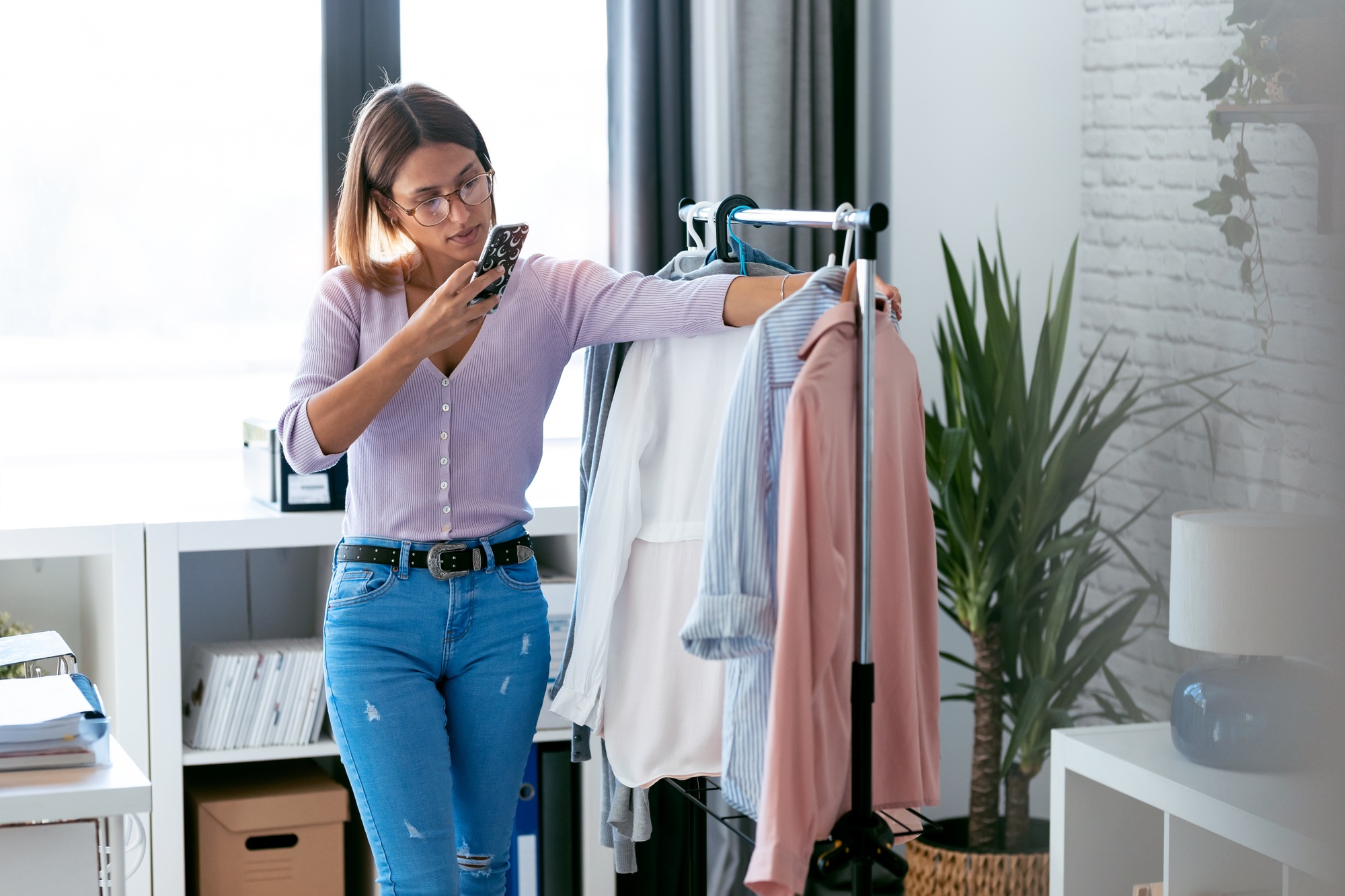 Beauty woman looking and choosing outfit while taking photos with her smart phone at home wardrobe.