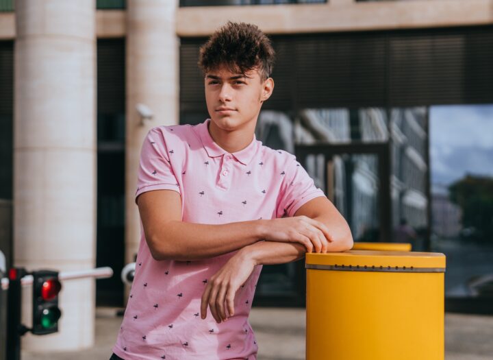 young man in pink polo shirt leans casually against yellow post. He gazes into distance with thought