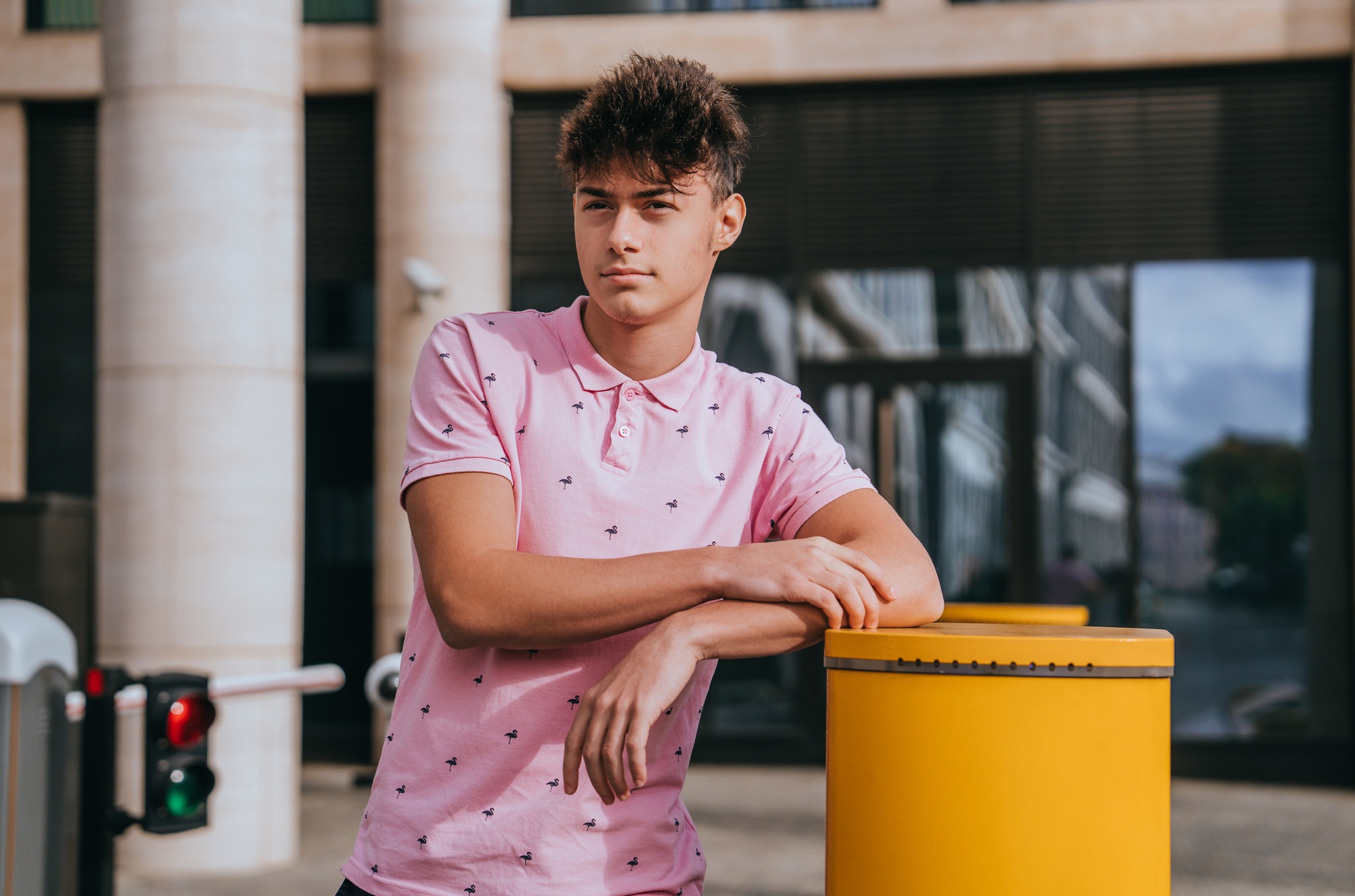 young man in pink polo shirt leans casually against yellow post. He gazes into distance with thought