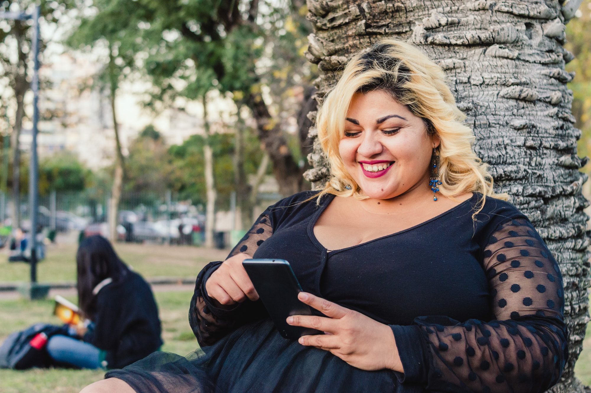 young plus size woman sitting on the grass in the park smiling and checking the phone